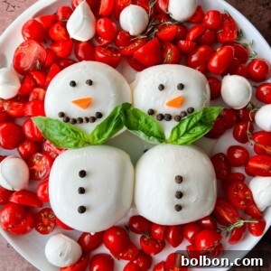 A plate of cherry tomatoes, mozzarella balls, and herbs, arranged with two Burrata Snowman figures made of mozzarella, decorated with peppercorn buttons, carrot noses, and basil leaf scarves.