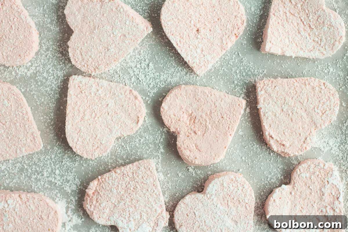 Several heart-shaped homemade strawberry marshmallows, some plain, some dipped in dark chocolate and topped with colorful sprinkles.