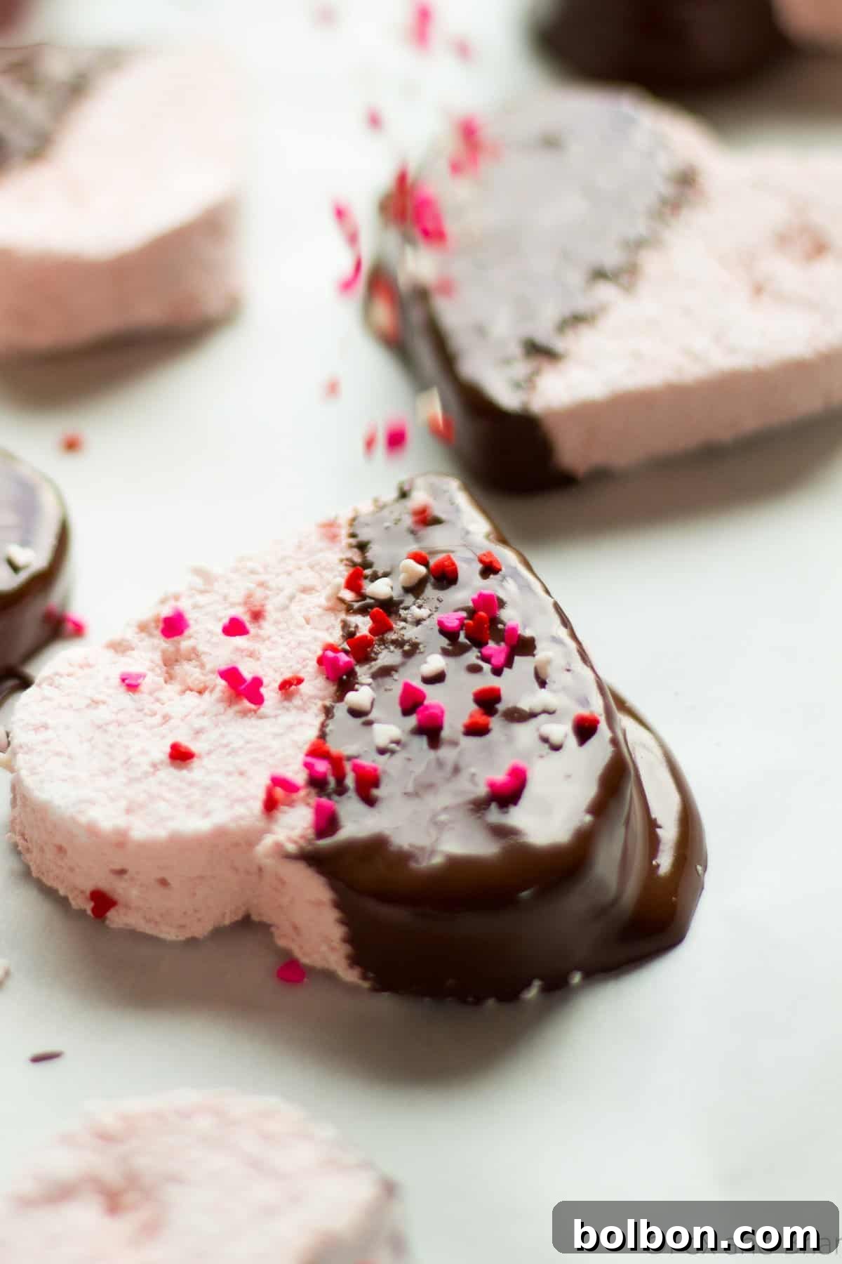 A heart-shaped homemade strawberry marshmallow, half-dipped in rich dark chocolate and generously covered in festive sprinkles, ready for gifting or enjoying.