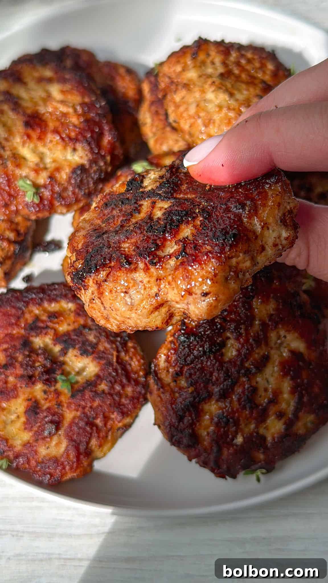 A hand holding a juicy, grilled burger patty with a golden-brown, slightly charred exterior. Several similar patties are arranged on a white plate below, with visible herbs and a glossy finish. The scene is lit by natural sunlight.