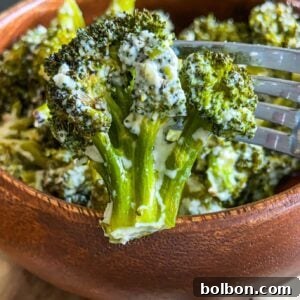 A close-up of Creamy Baked Broccoli florets coated in a luscious white sauce, with a fork lifting one piece above a wooden bowl.