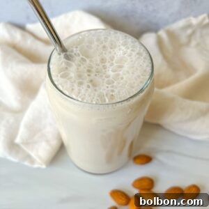 A glass of frothy almond milk made from a homemade almond milk recipe sits on a white surface, surrounded by whole almonds and a cream-colored cloth in the background.