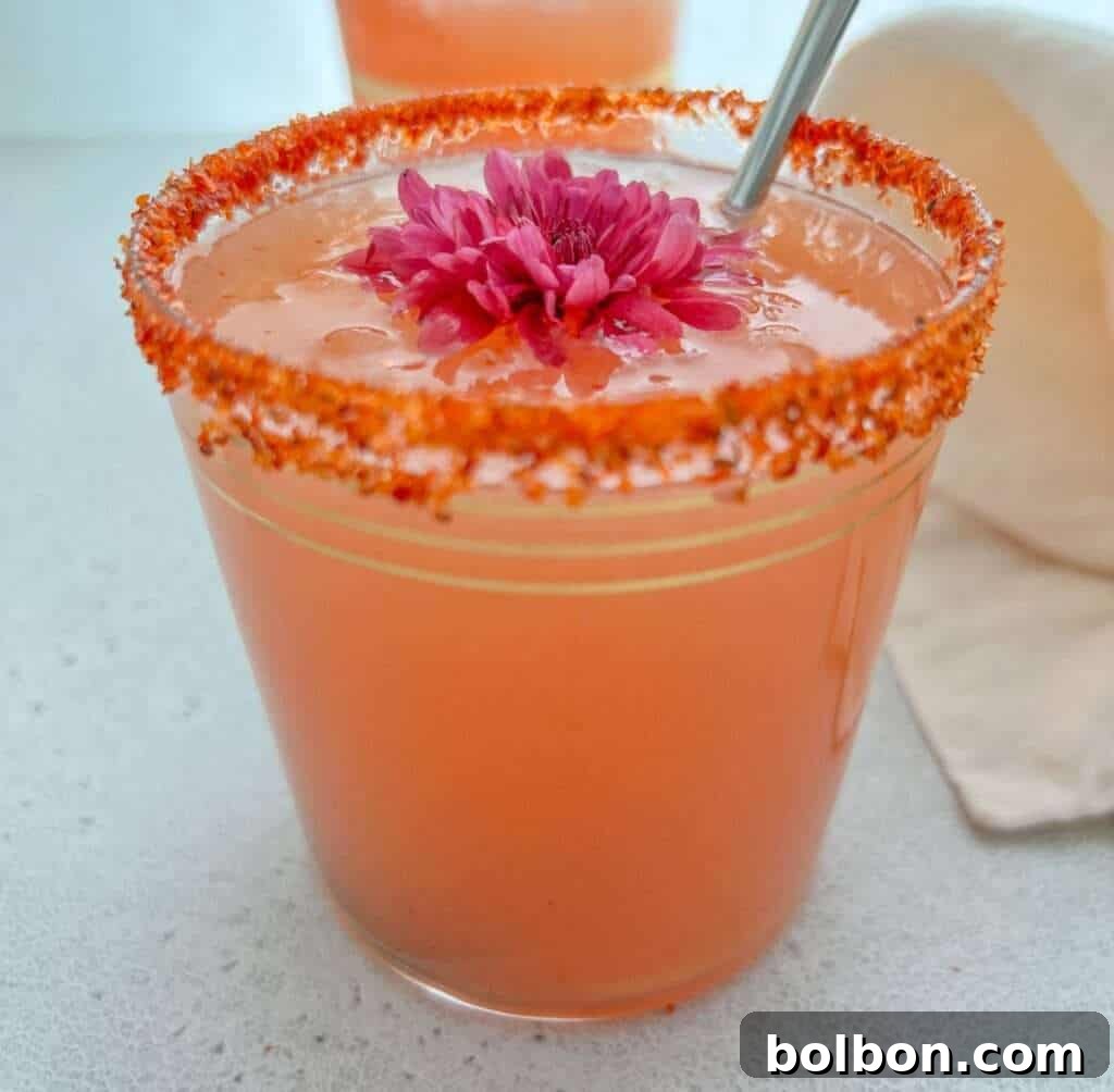 Pink Guava Margarita Magic 2 A vibrant pink guava margarita in a glass with a chili-lime rim, adorned with a tropical flower and a metal straw. The glass sits on a light surface with a beige napkin, evoking a holiday mood perfect for a spicy guava margarita recipe.