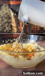 A hand mixer whips together eggs, sugar, and butter in a clear glass bowl, preparing the base for delicious Apple Cookies (Scones), while sunlight streams through a window in the background.