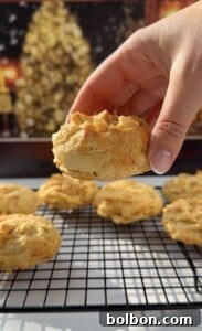 A hand holds a freshly baked Apple Cookies (Scones) biscuit above a cooling rack with more biscuits. A blurred, festive background with warm lights and a decorated tree sets the cozy holiday scene.