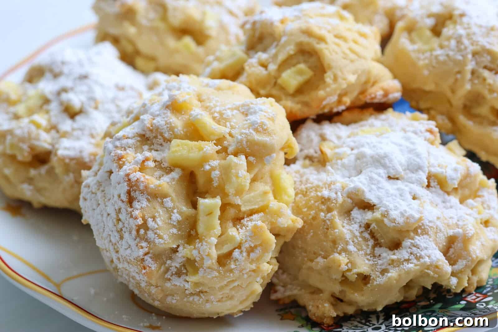 Close-up of several golden Apple Cookies (Scones) sprinkled with powdered sugar, arranged on a decorative plate with a colorful pattern in the corner. The cookies have a chunky texture and visible pieces of apple.