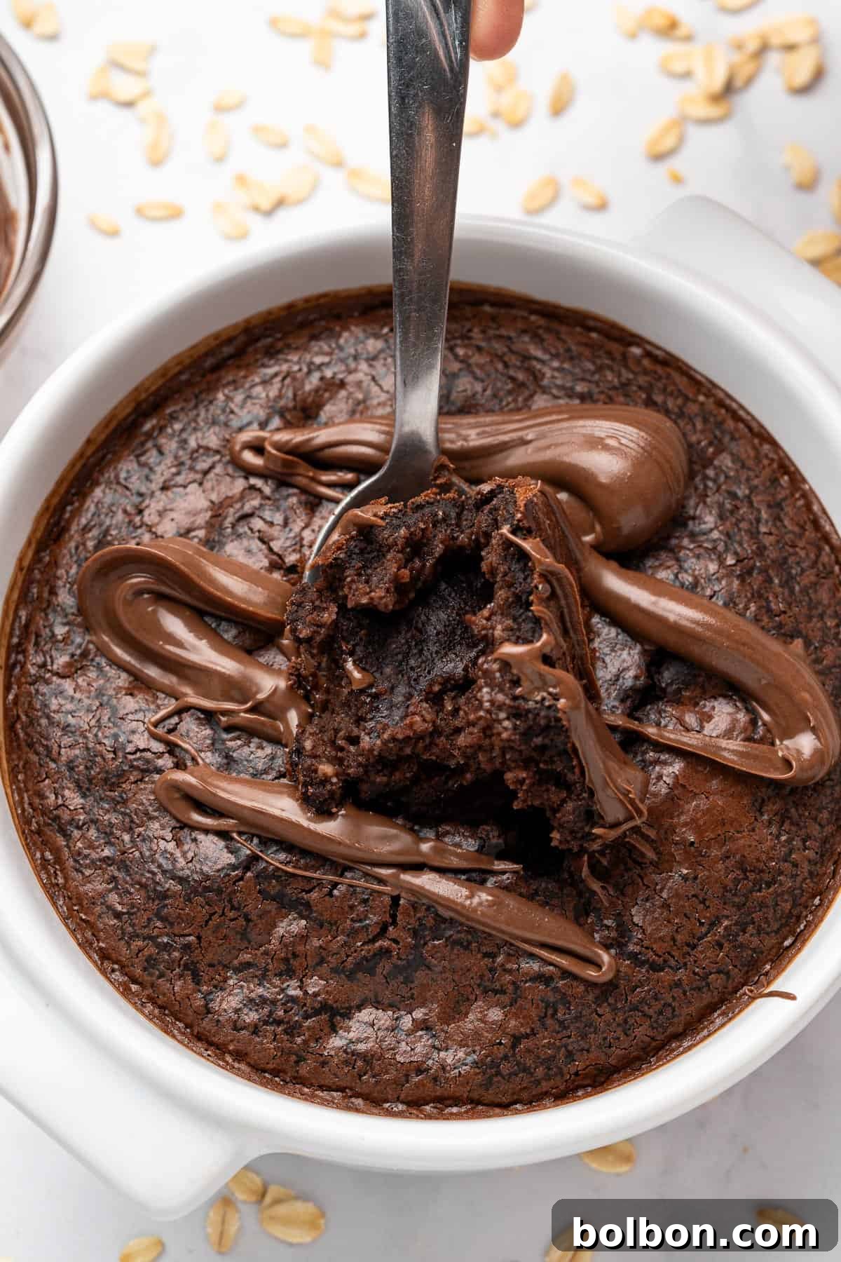 Fudgy Chocolate Oat Bar 2 A spoon taking a bite out of a chocolate dessert in a white dish, topped with swirls of melted chocolate. Oats are scattered around the dish on a light surface.