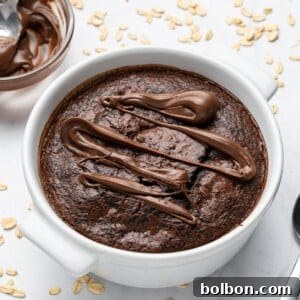A baked oatmeal dish topped with drizzled chocolate sits in a white bowl. A spoon rests beside it on a white surface scattered with oats. A bowl of chocolate spread is nearby.