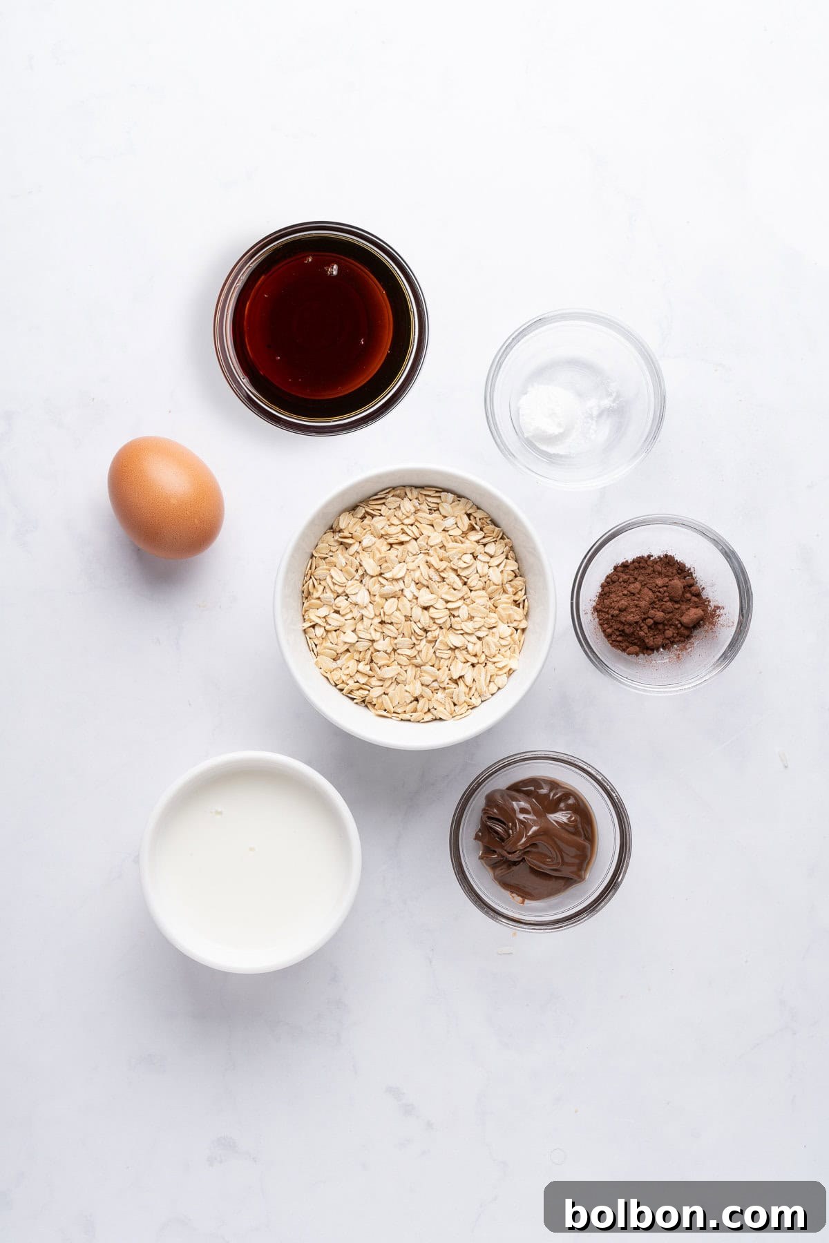 Fudgy Chocolate Oat Bar 3 A flat lay of baking ingredients on a white surface. Includes a bowl of oats, a glass of milk, an egg, small bowls of cocoa powder, baking soda, chocolate spread, and a bowl of maple syrup.