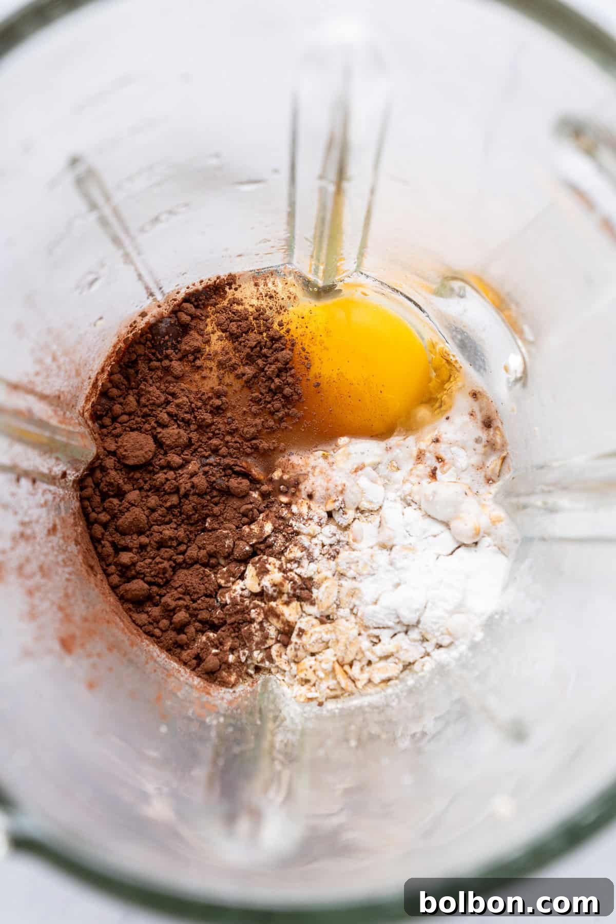 Fudgy Chocolate Oat Bar 4 Top view of a blender containing ingredients for baking, including cocoa powder, an egg, flour, oats, and a liquid. The ingredients are placed in different sections, ready to be blended.