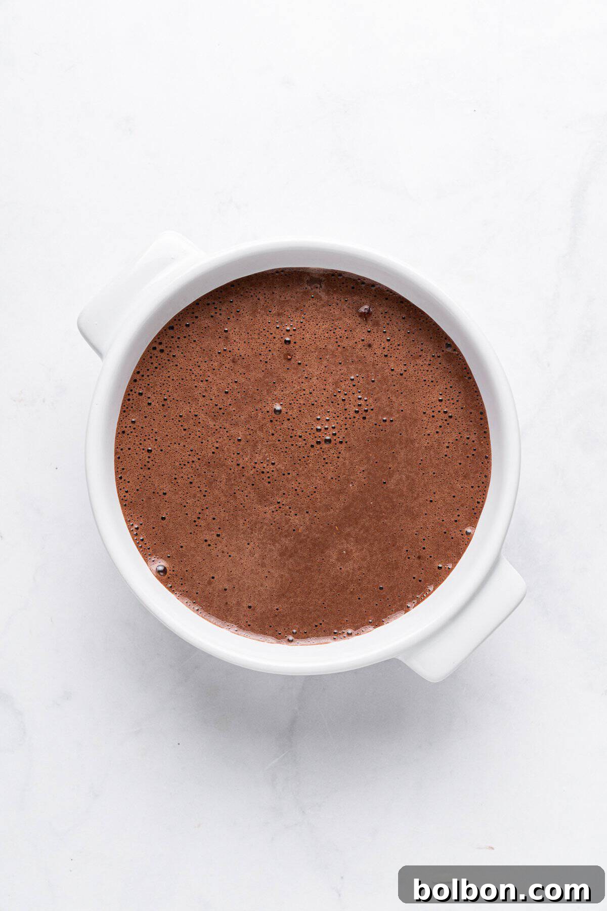 Fudgy Chocolate Oat Bar 6 A white ceramic bowl filled with smooth, rich chocolate batter on a light marble surface.