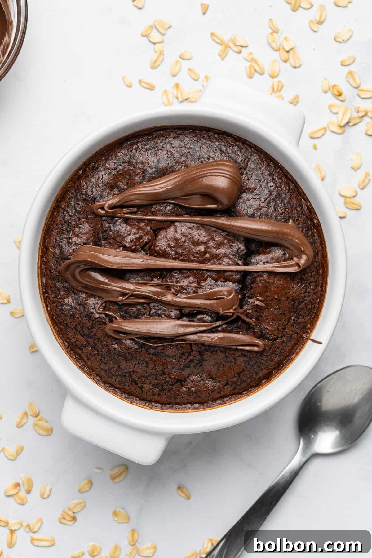 Fudgy Chocolate Oat Bar 7 A round white bowl filled with a baked chocolate oat dessert, topped with drizzles of smooth chocolate. A spoon lies beside the bowl on a white surface, scattered with oats.