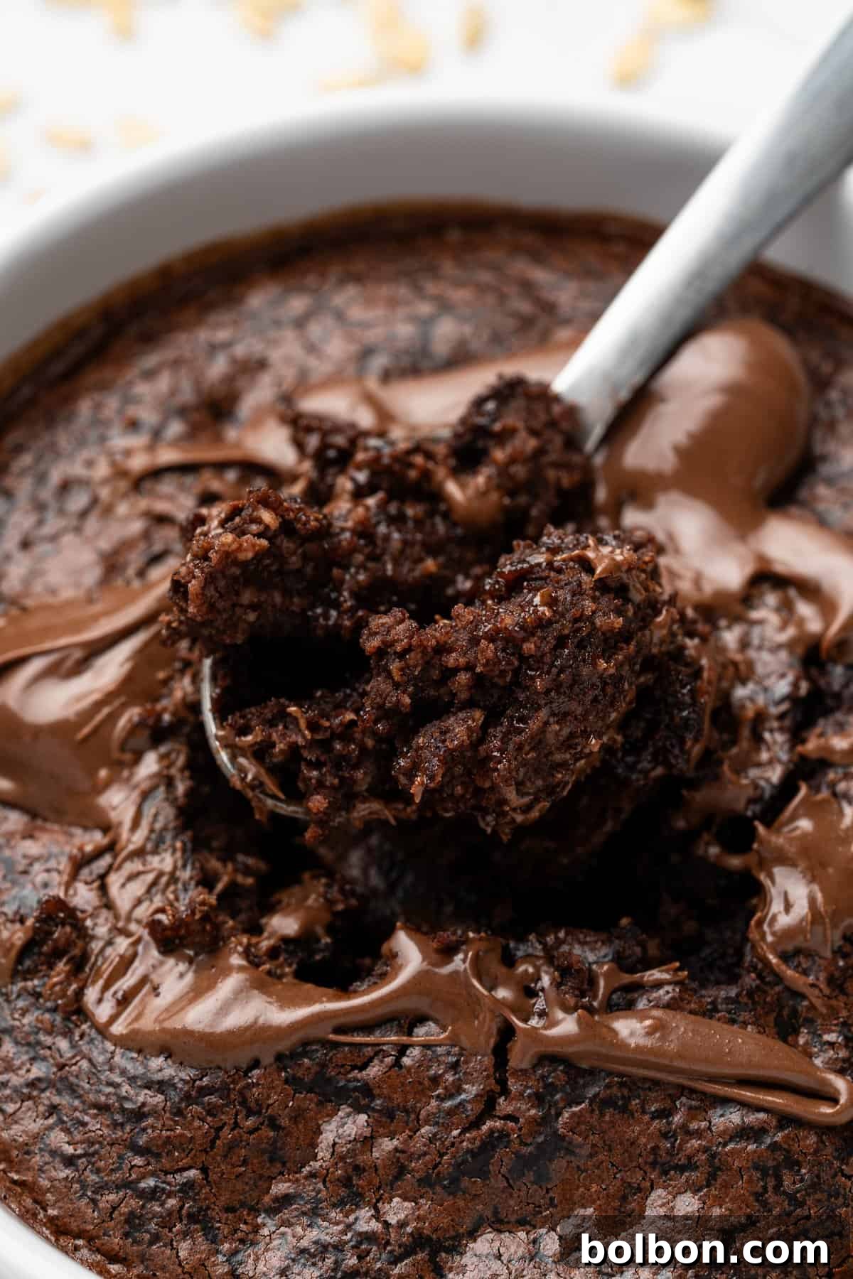 Fudgy Chocolate Oat Bar 8 A close-up of a spoon scooping a portion of gooey, baked chocolate dessert from a white dish. The surface is rich and textured, with melted chocolate swirls. The background is blurred with a hint of scattered ingredients.