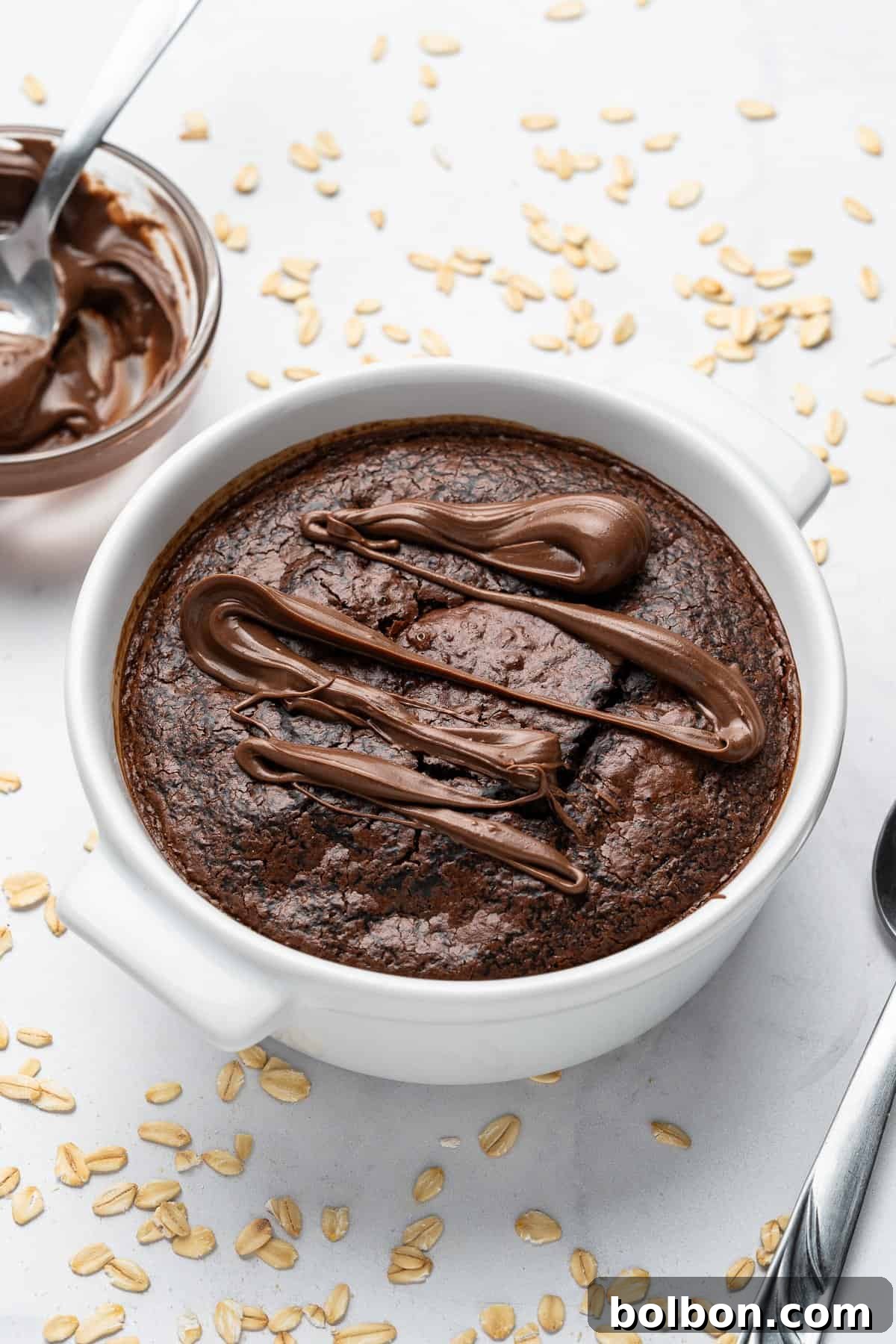 Fudgy Chocolate Oat Bar 9 A baked oatmeal dish topped with drizzled chocolate sits in a white bowl. A spoon rests beside it on a white surface scattered with oats. A bowl of chocolate spread is nearby.