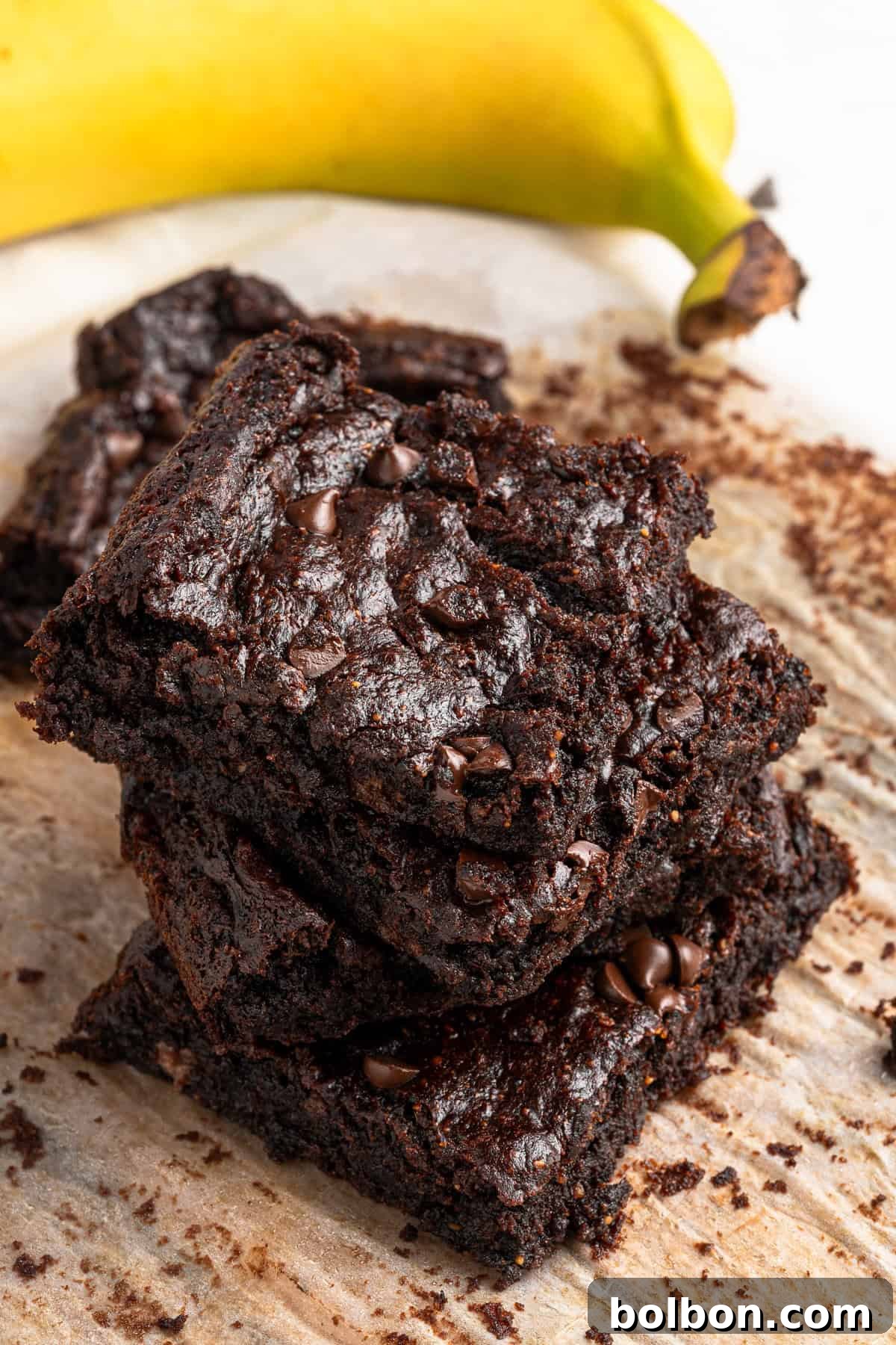 A stack of rich, fudgy chocolate brownies with visible chocolate chips on top, perfectly arranged on a piece of parchment paper. A vibrant, ripe banana rests in the background, hinting at the key ingredient and adding a pop of natural color to the scene.