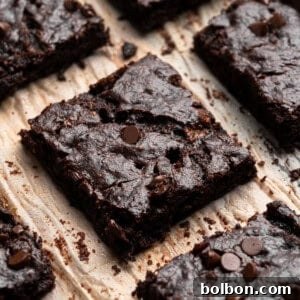 Close-up of several rich, chocolate brownies topped with chocolate chips, arranged on parchment paper. The brownies have a dense, fudgy texture, and the chocolate chips add a touch of shine.