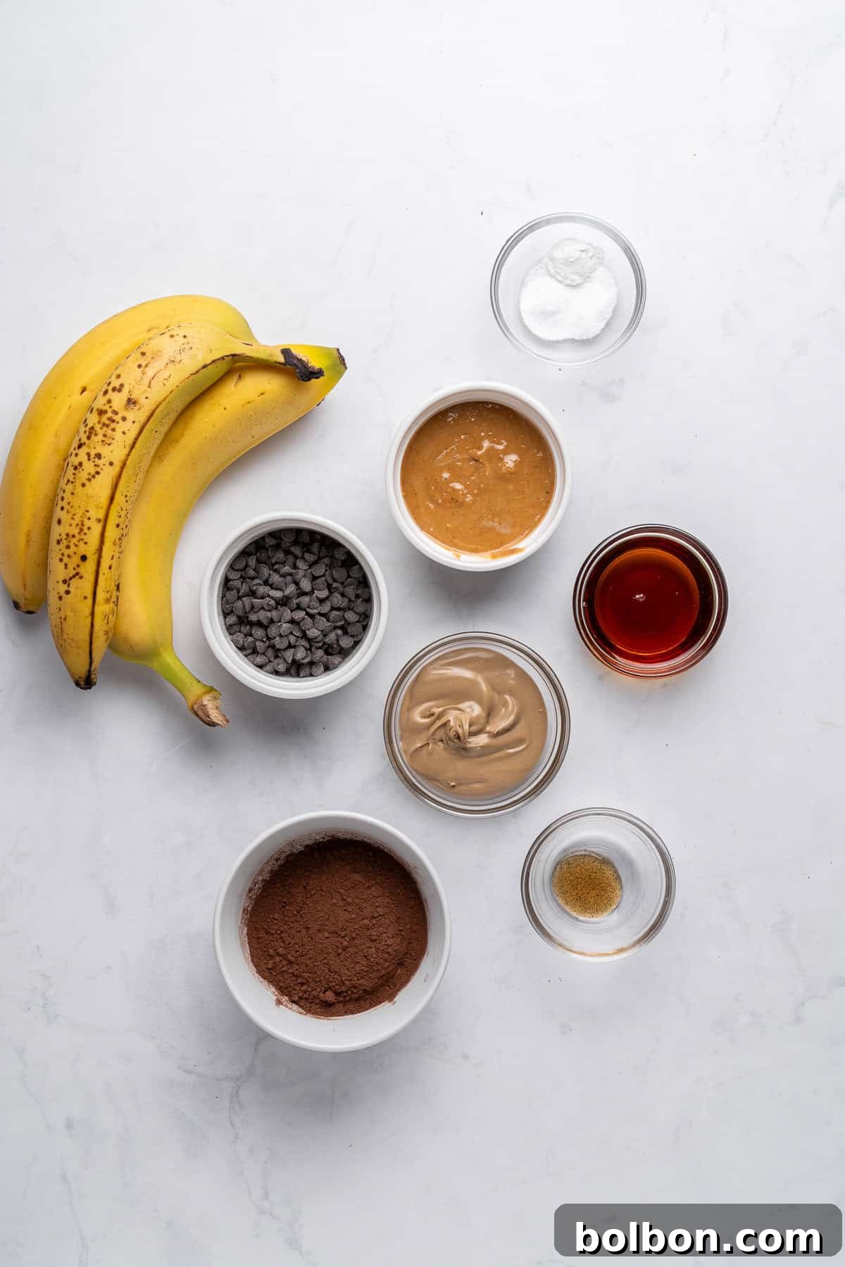 Ingredients for a baking recipe neatly arranged on a sleek marble surface: two ripe bananas, small bowls of rich chocolate chips, unsweetened cocoa powder, creamy almond butter, natural peanut butter, a glistening glass of maple syrup, and tiny bowls holding essential baking soda and aromatic vanilla extract, all ready for mixing.