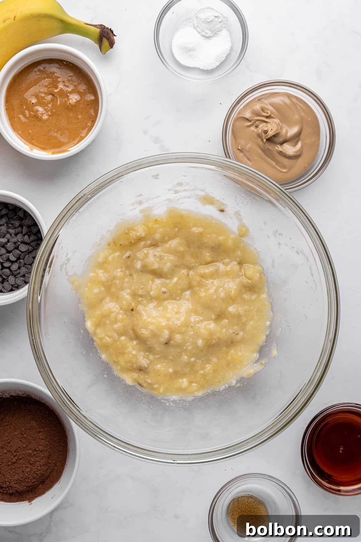 A clear glass bowl containing perfectly mashed bananas sits prominently on a marble surface. Surrounding it are various baking ingredients in small bowls: creamy peanut butter, glistening chocolate chips, dark cocoa powder, a whole ripe banana, a sprinkle of baking soda, a dash of salt, a few drops of vanilla extract, and a measure of brown sugar, all contributing to a wholesome recipe.
