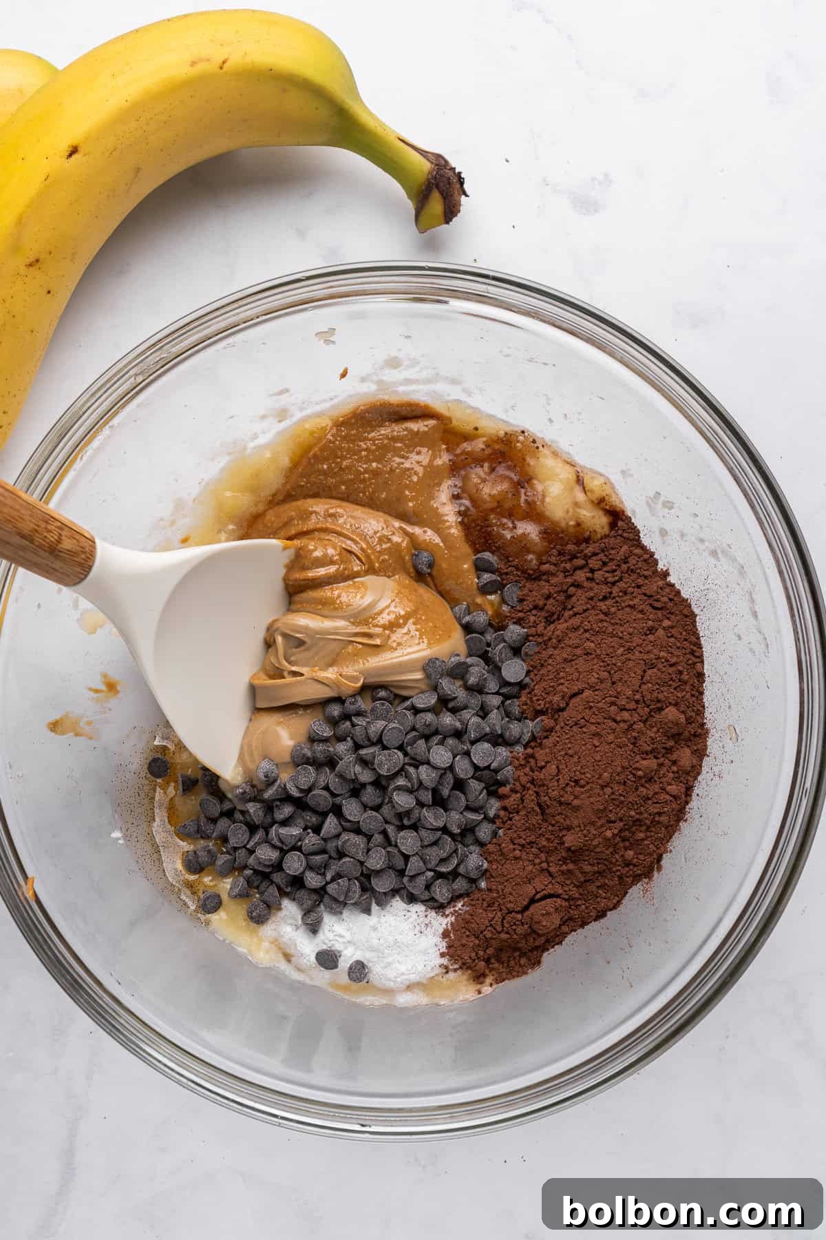 A glass bowl holds the beginnings of a delicious baking creation: a mixture of mashed banana, creamy peanut butter, rich chocolate chips, and dark cocoa powder. A white spatula is actively stirring the ingredients, ensuring they are well combined. Beside the bowl, on a pristine marble surface, rests a whole ripe banana, awaiting its turn or simply adding to the aesthetic.
