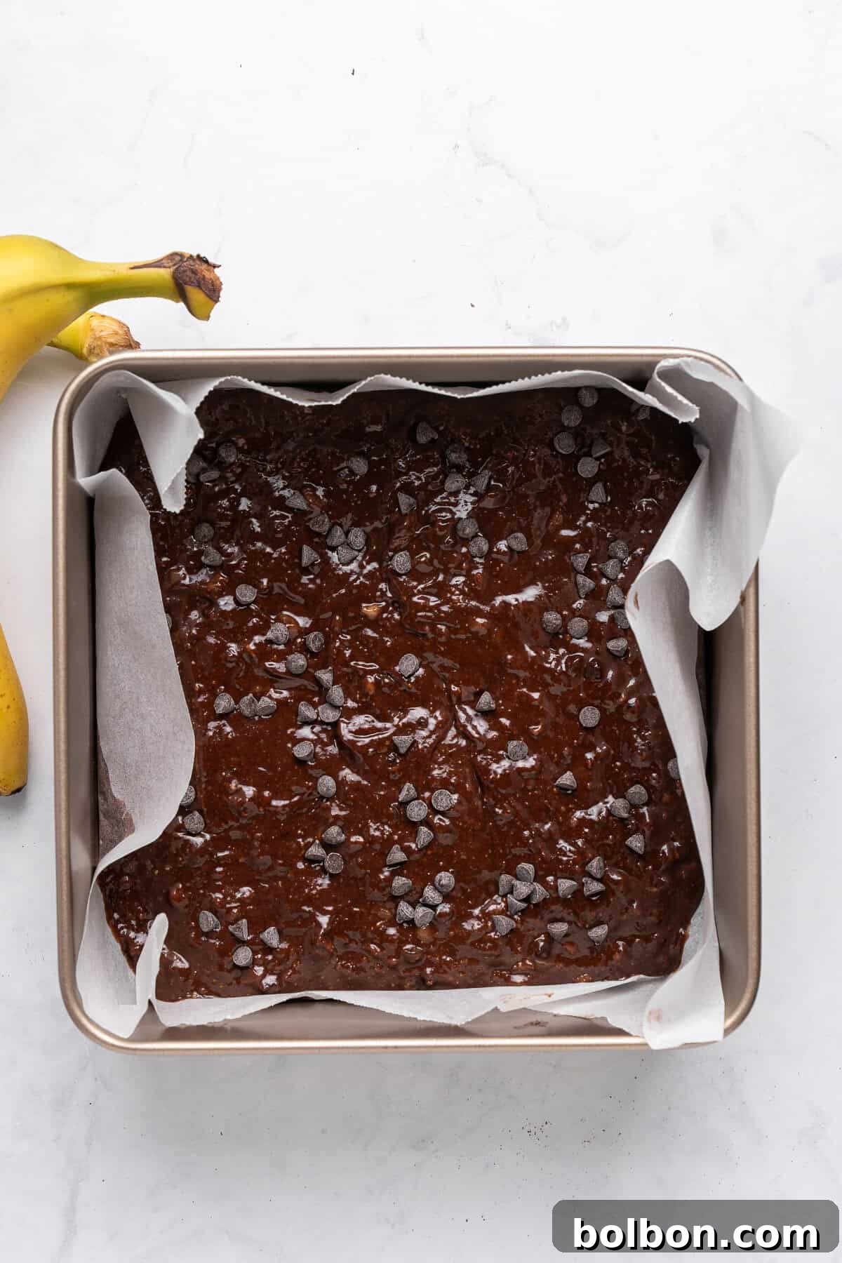 A square baking pan, neatly lined with parchment paper, is filled with raw, rich chocolate brownie batter. The top of the batter is generously sprinkled with chocolate chips, promising a decadent treat. Two ripe bananas are partially visible on the side, resting on a clean white marble surface, suggesting the key ingredient.