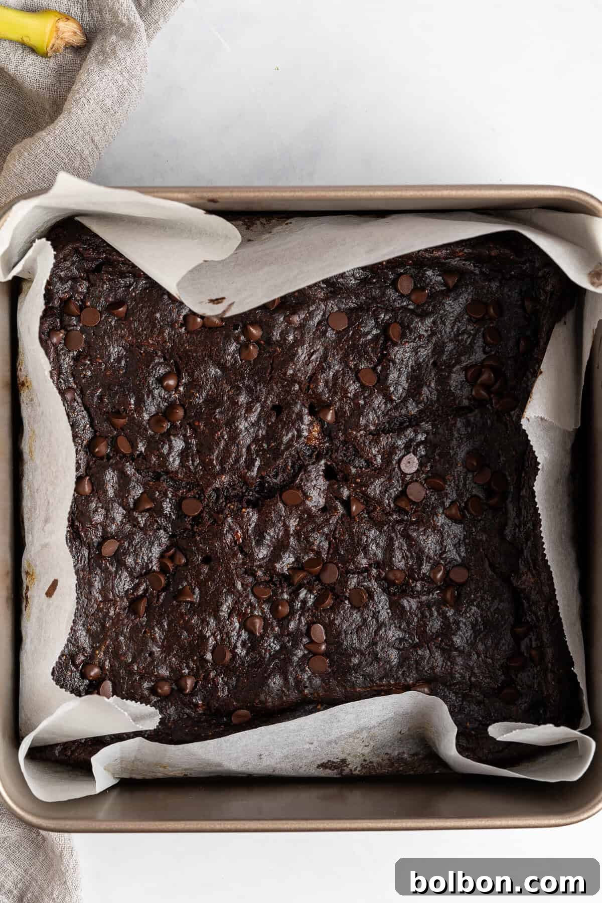 A square baking pan, lined with parchment paper, holds a batch of freshly baked chocolate brownies. The brownies are dark, glossy, and beautifully topped with melted chocolate chips. Their surface appears rich, moist, and perfectly set, indicating a fudgy interior. A beige fabric is partially visible on the side, adding a soft background element.