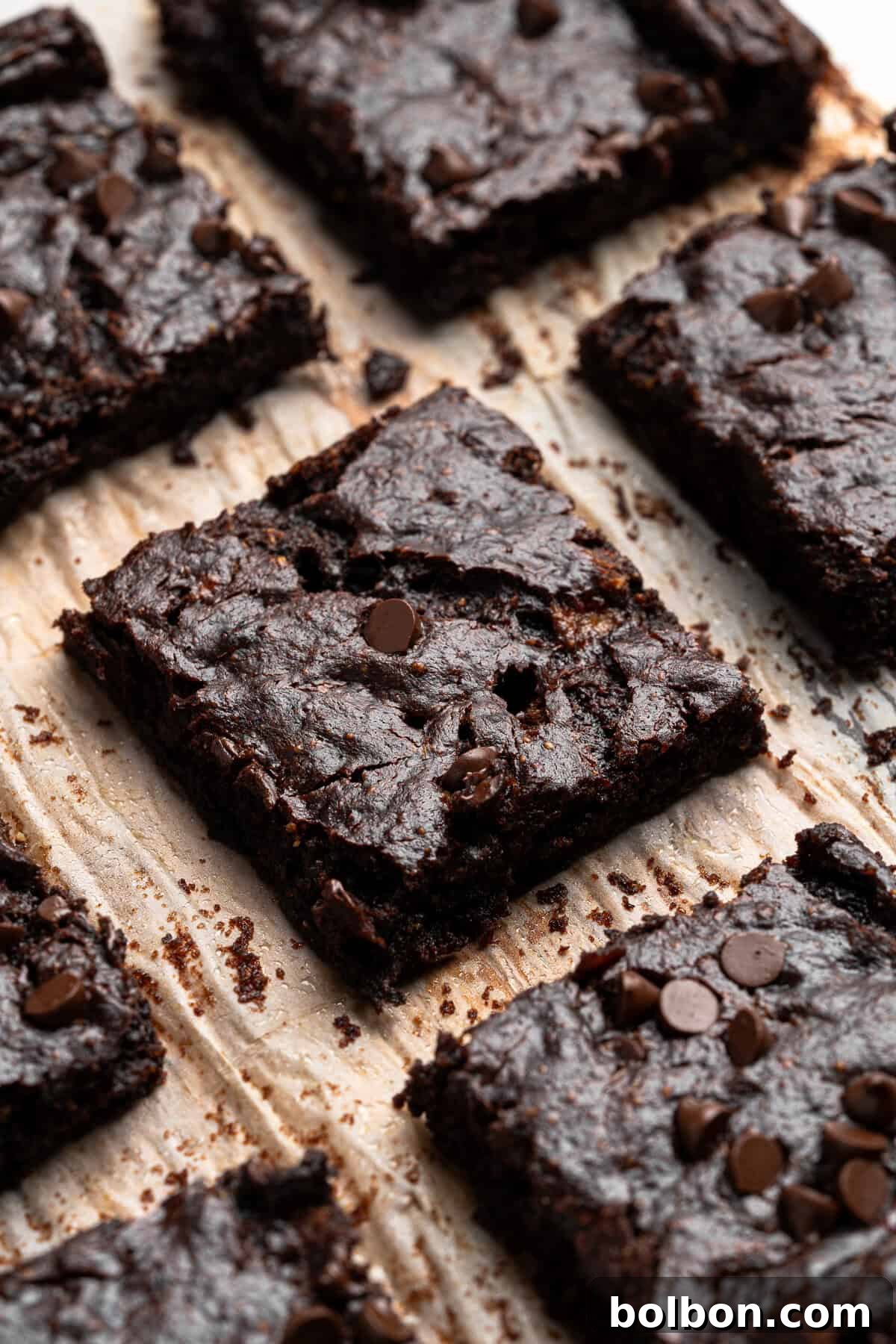 A close-up view of several rich, dark chocolate brownies, generously topped with chocolate chips. They are arranged on a piece of parchment paper, showcasing their dense, fudgy texture and glossy surface. The melted chocolate chips add a touch of sparkle and allure.