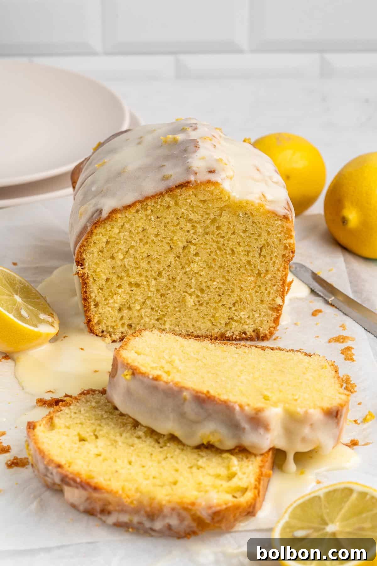 Zesty Whole Lemon Blender Cake 2 Lemon loaf cake with lemon glaze, sliced on a marble surface. Two whole lemons and lemon slices are placed nearby. A knife and stacked plates are visible in the background.