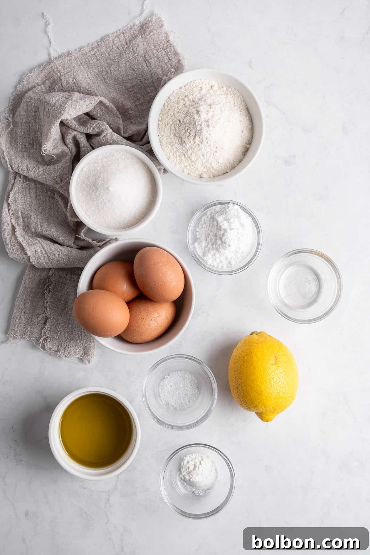 Zesty Whole Lemon Blender Cake 4 A flat lay of baking ingredients on a marble surface, including flour, sugar, eggs, a lemon, olive oil, baking powder, salt, and water. A gray cloth napkin is placed alongside the ingredients.