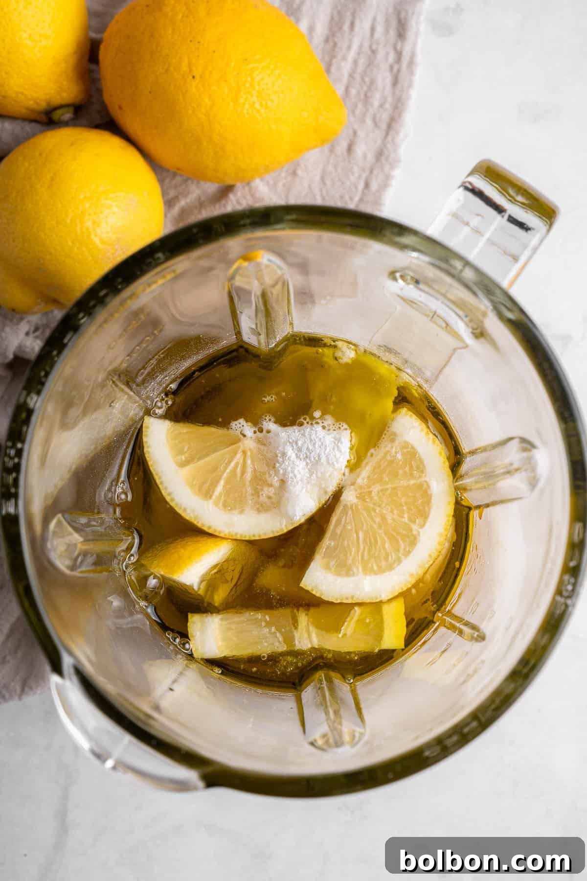 Zesty Whole Lemon Blender Cake 5 Top view of a blender containing lemon slices, olive oil, and a sprinkle of salt, placed on a light-colored surface. Three whole lemons rest beside the blender on a textured cloth.