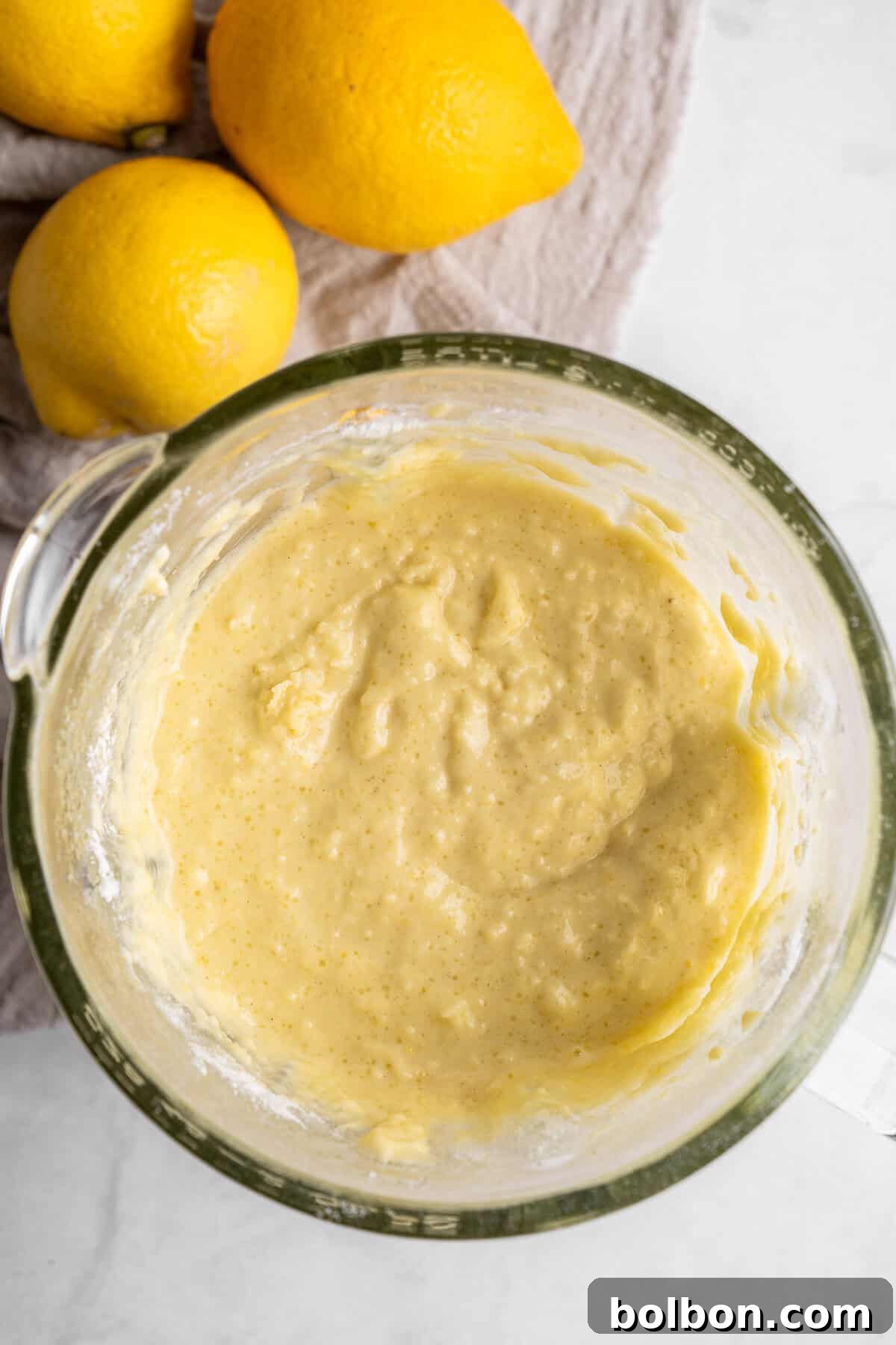 Zesty Whole Lemon Blender Cake 6 A glass mixing bowl filled with creamy lemon batter on a white countertop. Three whole lemons lie on a beige cloth beside the bowl.
