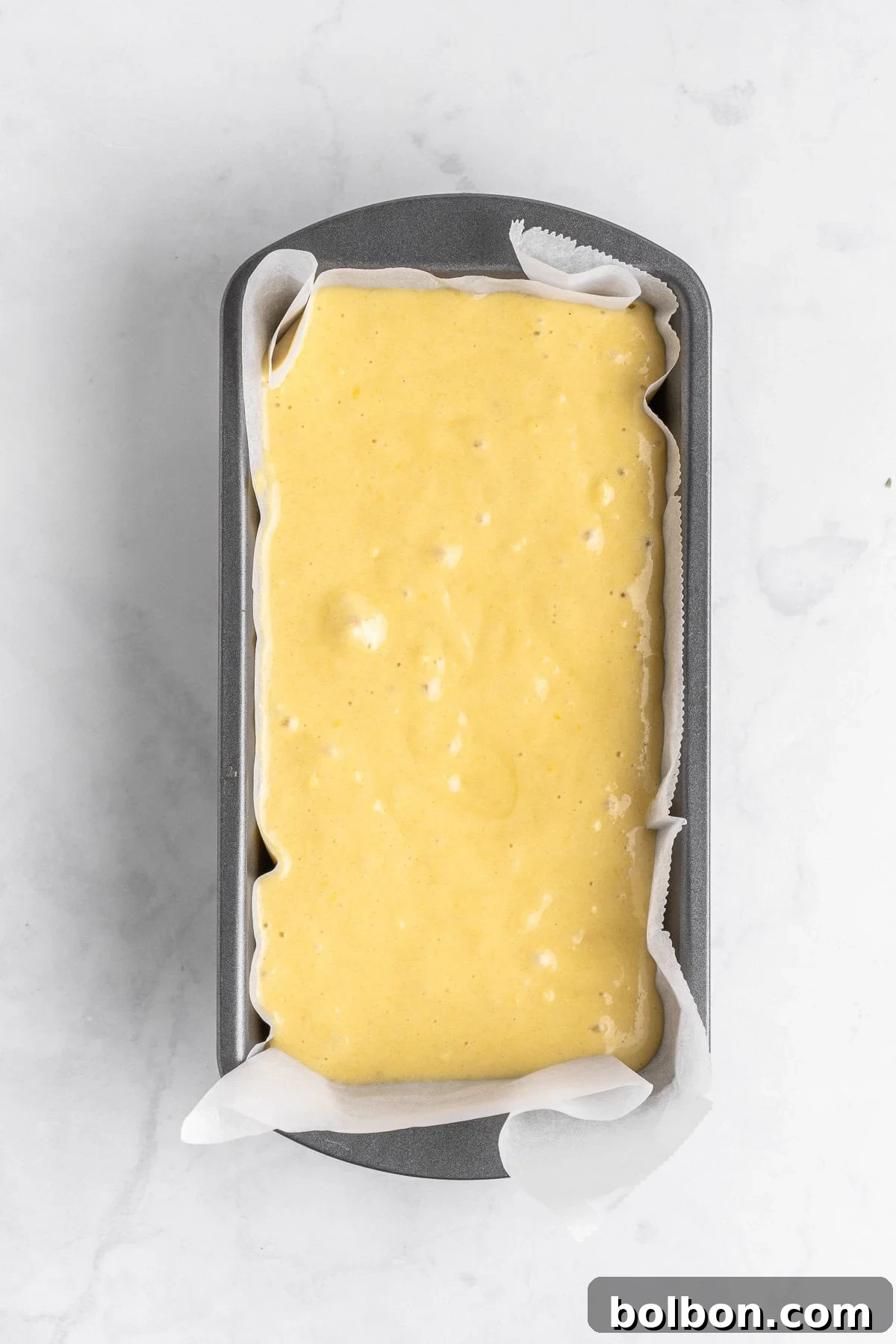 Zesty Whole Lemon Blender Cake 7 A loaf pan lined with parchment paper is filled with uncooked yellow batter, ready to be baked, set against a white marble background.
