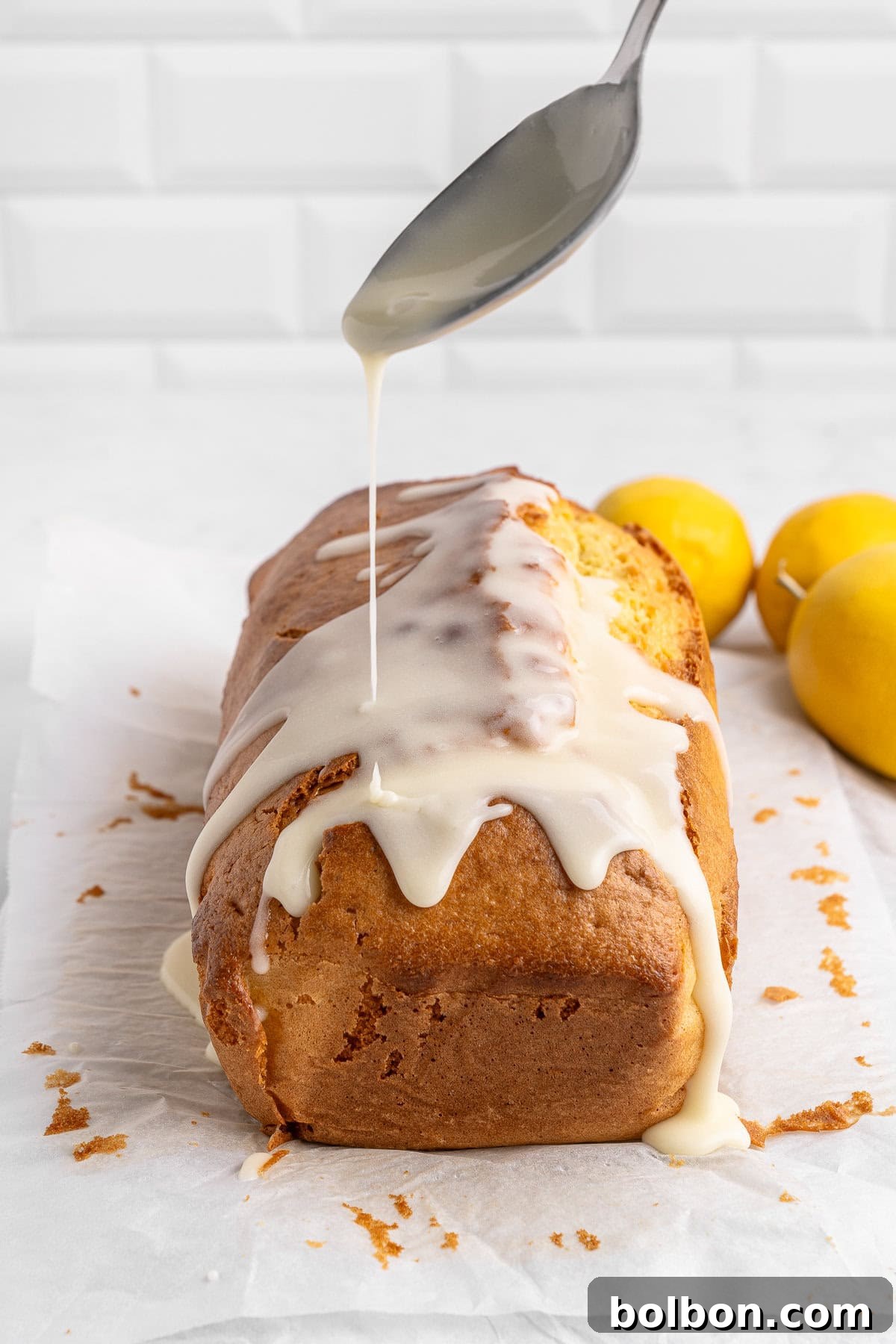 Zesty Whole Lemon Blender Cake 9 A loaf of lemon cake on parchment paper is being drizzled with icing from a spoon. Two whole lemons are placed in the background. The setting is a white kitchen counter with a tiled backsplash.