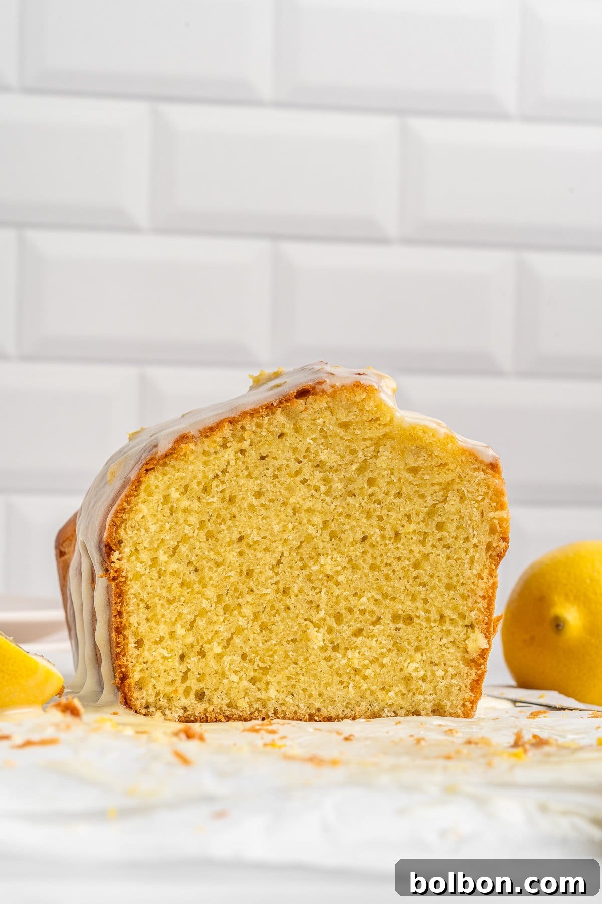 Zesty Whole Lemon Blender Cake 10 A sliced loaf of lemon drizzle cake with a light glaze on top is displayed against a white tiled background. Lemons are placed nearby, and crumbs are scattered around the cake.
