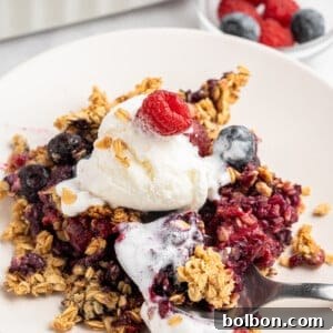 A serving of mixed berry crisp topped with a scoop of vanilla ice cream and garnished with a raspberry on a white plate. The crisp contains oats and various berries, with a fork resting on the plate. A dish of additional berries is in the background.