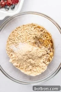 Summer Berry Crunch 8 A glass bowl filled with oats, almond flour, and brown sugar, ready for mixing. On the side, a dish with assorted berries adds a colorful contrast. The ingredients are set against a white background, suggesting preparation for a recipe.
