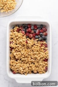 Summer Berry Crunch 9 A white baking dish with a partially covered layer of mixed berries, including raspberries and blueberries, topped with uncooked oat crumble mixture. There is a glass bowl with more oats in the top left corner.