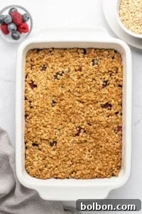 Summer Berry Crunch 10 A baked oatmeal dish in a white rectangular casserole dish on a marble surface. Next to it are a bowl of mixed berries and a bowl of oats. A folded beige cloth is placed nearby.