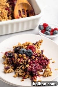 Summer Berry Crunch 11 A plate with a serving of berry crisp topped with granola and fresh blueberries and raspberries. In the background, a baking dish with more crisp and a small bowl of berries are visible.
