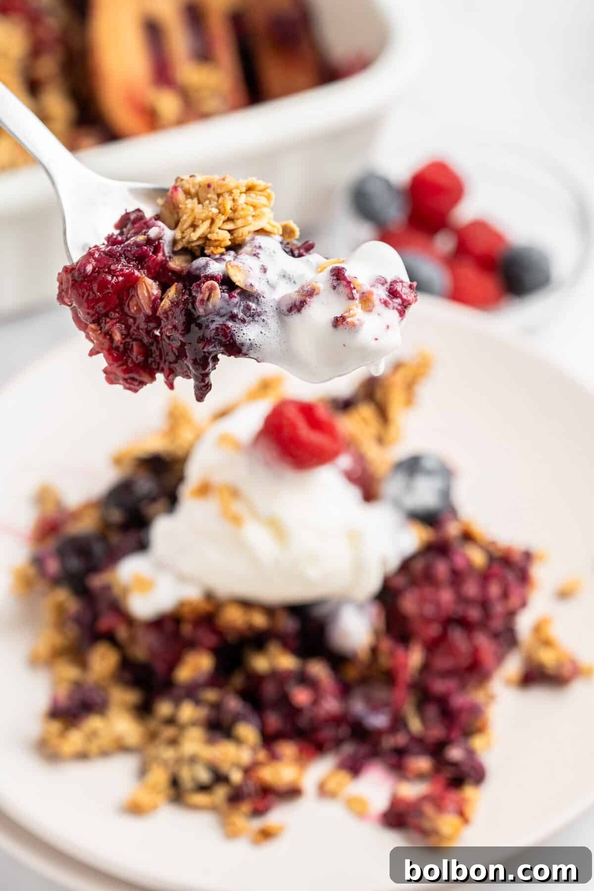 Summer Berry Crunch 3 A spoonful of the crisp with granola and a dollop of whipped cream is held above a plate. In the background, a dish with more berry crisp and a small bowl of fresh berries are visible.