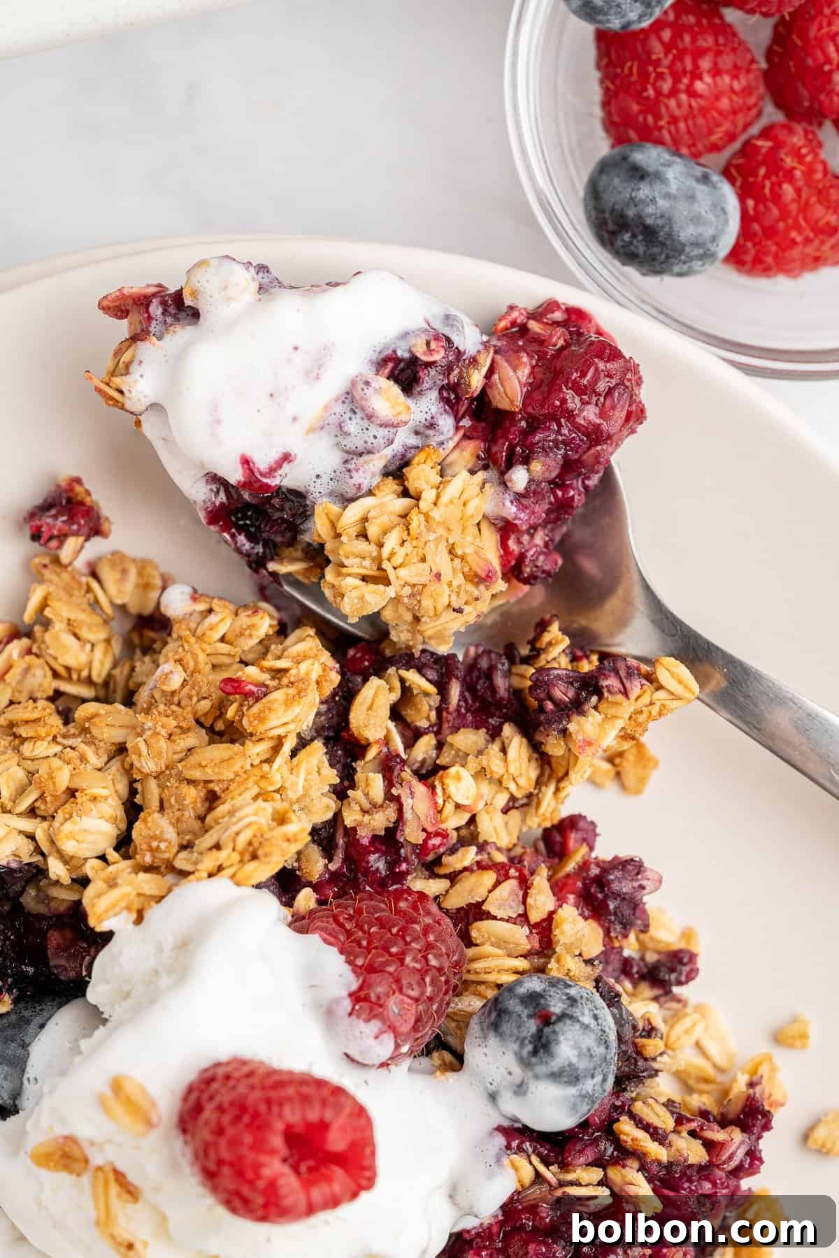 Summer Berry Crunch 4 A close-up of a breakfast dish featuring granola, fresh raspberries, and blueberries on a plate. A spoon with some yogurt and granola is beside it. A small bowl with more berries is in the background.