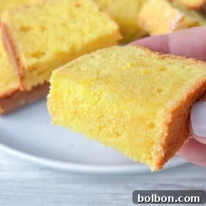 A hand holding a slice of moist yellow Clementine Cake with more slices stacked on a white plate in the background on a light wooden surface.