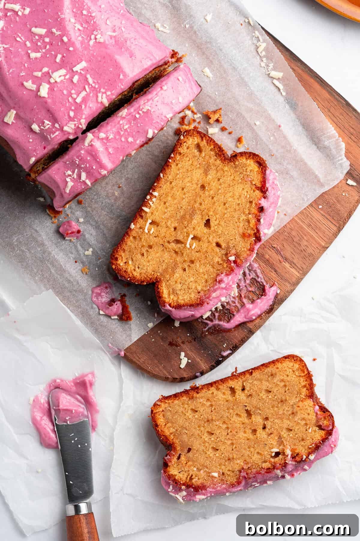 A beautifully sliced loaf cake with a luscious pink frosting rests on a rustic wooden board. Two perfect slices are thoughtfully placed beside it, hinting at the moist, fluffy interior. A knife, still adorned with a trace of the smooth frosting, lies on parchment paper nearby, while delicate white chocolate shavings are scattered as an elegant garnish.