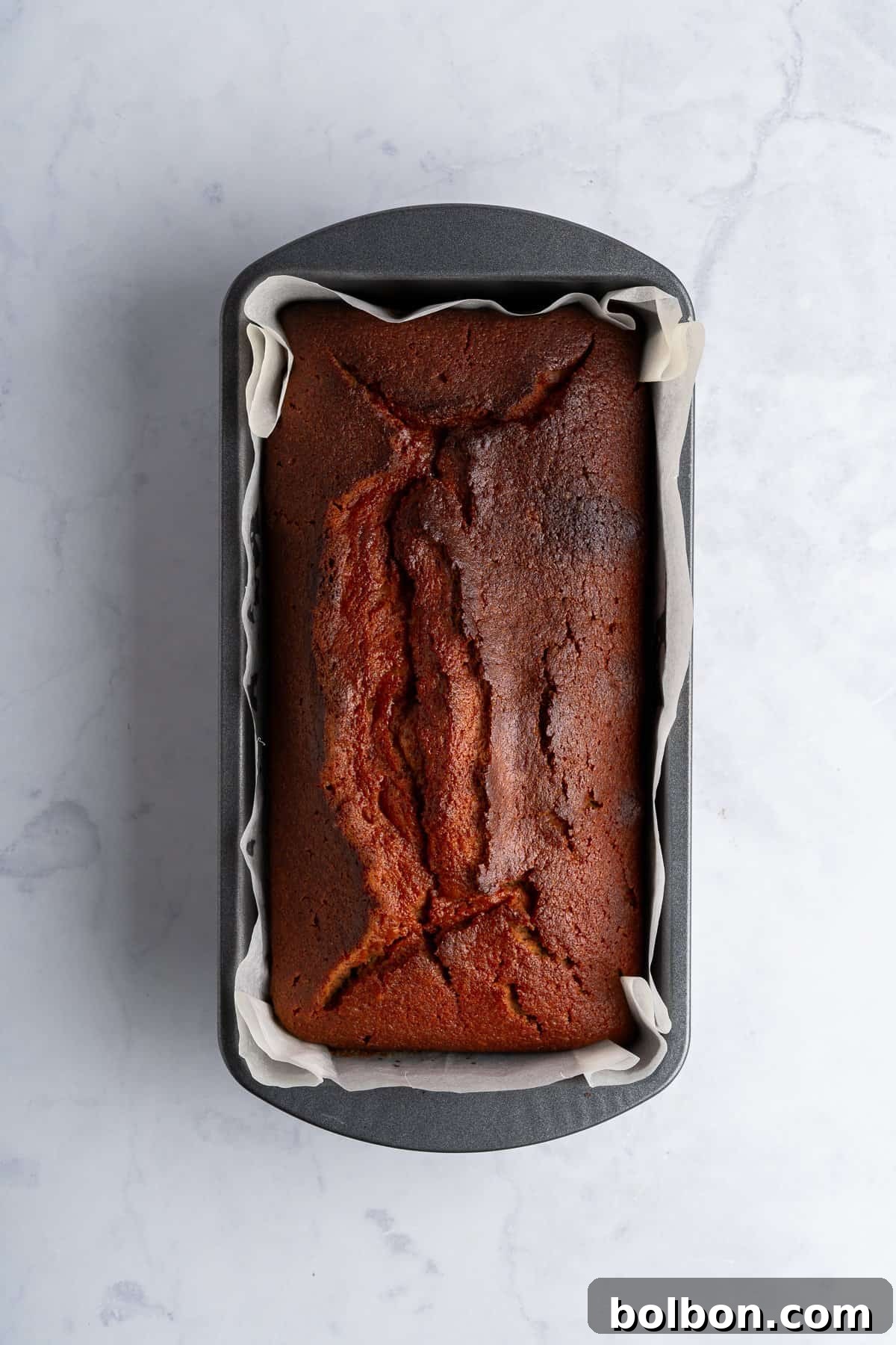 A freshly baked loaf of golden-brown banana bread rests perfectly in a rectangular metal pan, lined with parchment paper, on a light marble surface. The bread's top displays a beautiful golden hue with slight, natural cracks, indicating its readiness.