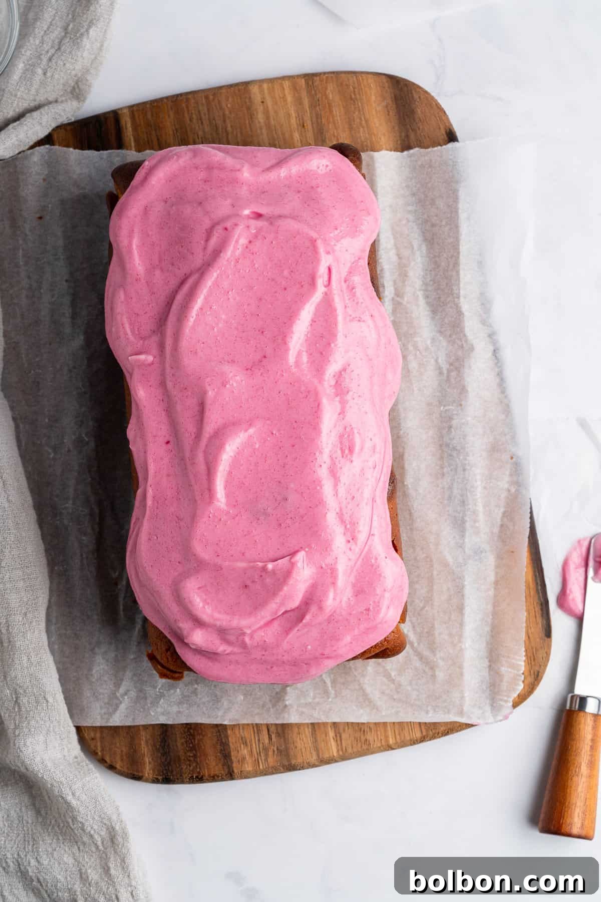 A beautifully decorated rectangular loaf cake, adorned with bright pink frosting, is presented on a wooden board, partially covered with parchment paper. A knife, still carrying traces of the creamy pink frosting, is carefully placed nearby on a pristine white surface, next to a casually draped gray cloth.