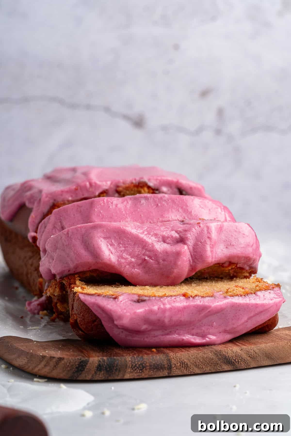 A delightful sliced loaf cake, crowned with a rich, vibrant pink frosting, is beautifully arranged on a wooden board. The cake's moist interior is visible through the slices, and the frosting is generously and smoothly spread, hinting at a creamy, luxurious texture. The entire setup is presented against a soft, light gray background.
