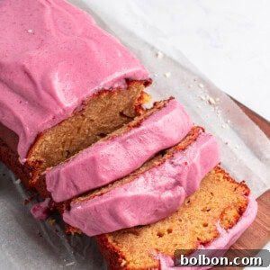 A vibrant loaf of cake generously coated with bright pink frosting, partially sliced, rests on a wooden board. Delicate white chocolate shavings are scattered in the background on a light surface, adding an elegant touch.