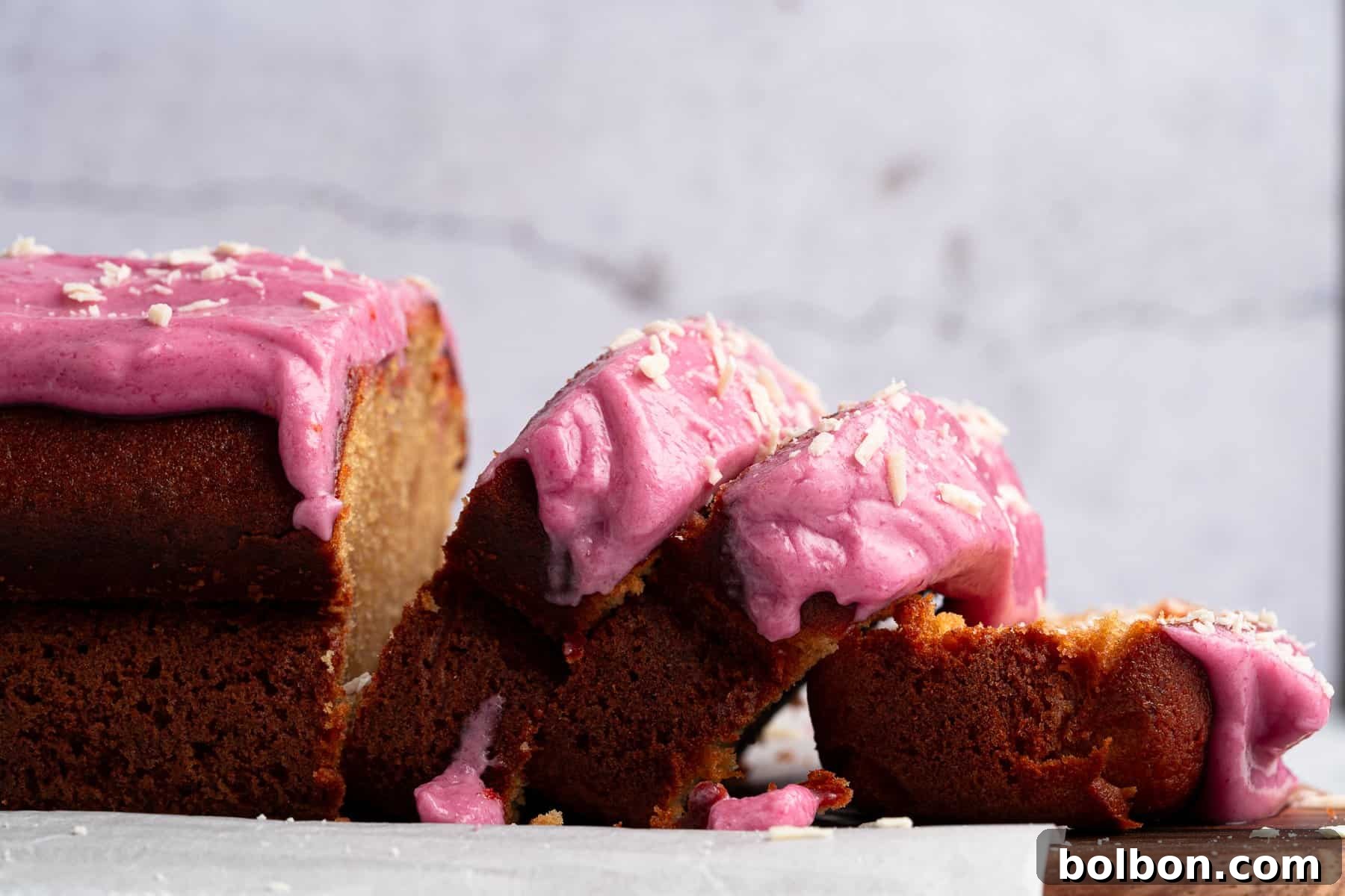 A rustic wooden board holds a partly sliced loaf cake, richly coated with vibrant pink frosting, revealing the cake's moist and tender interior. Scattered crumbs and remnants of the creamy frosting surround the cake on a light-colored surface, creating an inviting visual contrast against a softly blurred, natural background.