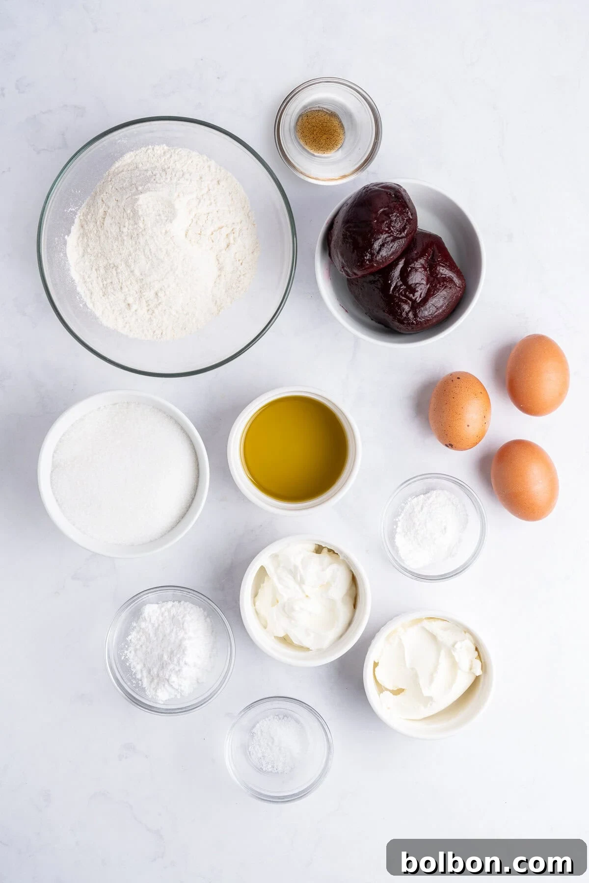 A top-down view showcases an array of fresh baking ingredients, meticulously arranged in individual bowls on a pristine white surface. Included are all-purpose flour, granulated sugar, two vibrant cooked beets, farm-fresh eggs, smooth olive oil, baking powder, baking soda, creamy yogurt, fine salt, and aromatic vanilla extract, all poised for the blending process.