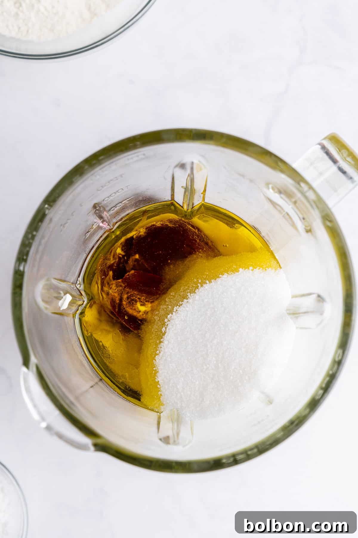 From a top-down perspective, a clear blender pitcher sits on a white surface, holding a base of oil, sugar, and vanilla extract. The translucent glass perfectly reveals the distinct layers of ingredients, awaiting the next step of mixing to transform into a smooth batter.