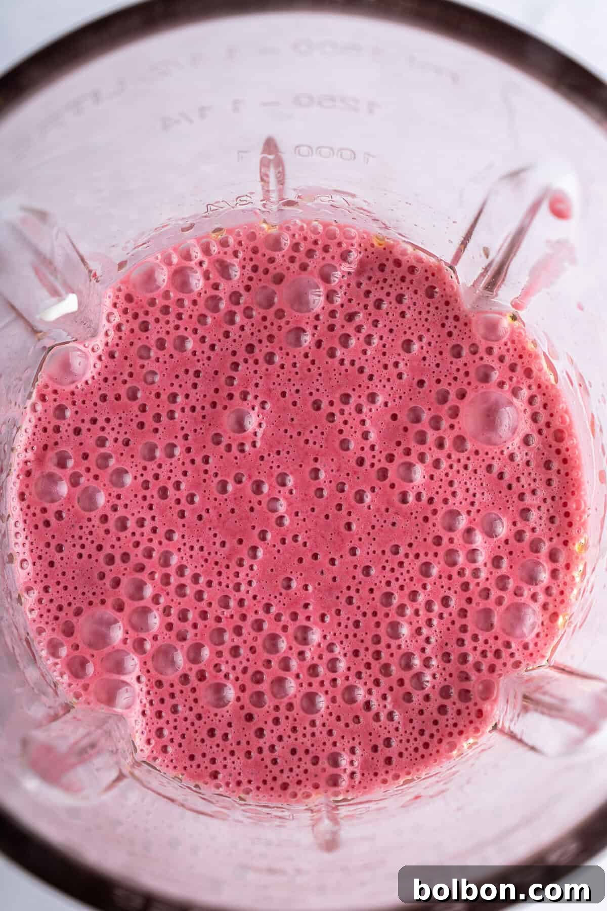 A blender pitcher, filled with a frothy, vibrant pink smoothie, showcases a delightful bubbly texture on its surface. The clear glass of the blender jar reveals visible measurement markings on its side, indicating the volume of the perfectly blended mixture.